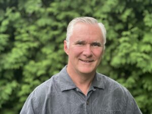 Steve Horner face on. Steve has a grey shirt on, white hair, and is standing in front of a hedge. This is a typical head shot sort of image.